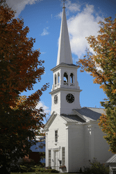 An old white chapel is bordered by two trees in the fall.