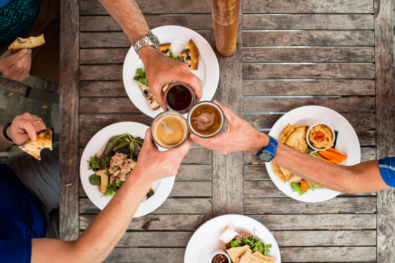 Seen from above, three people toast beers poured in glasses with food on the table.