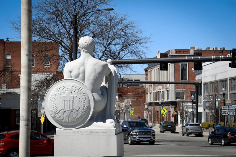 Seen from behind, a granite statue of a person with a shield, overlooks a road intersection in a small rural town.