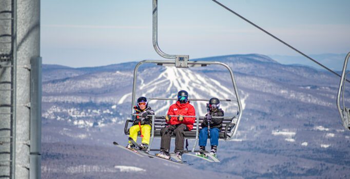 Three people on a chairlift with mountains visible behind on a sunny day.