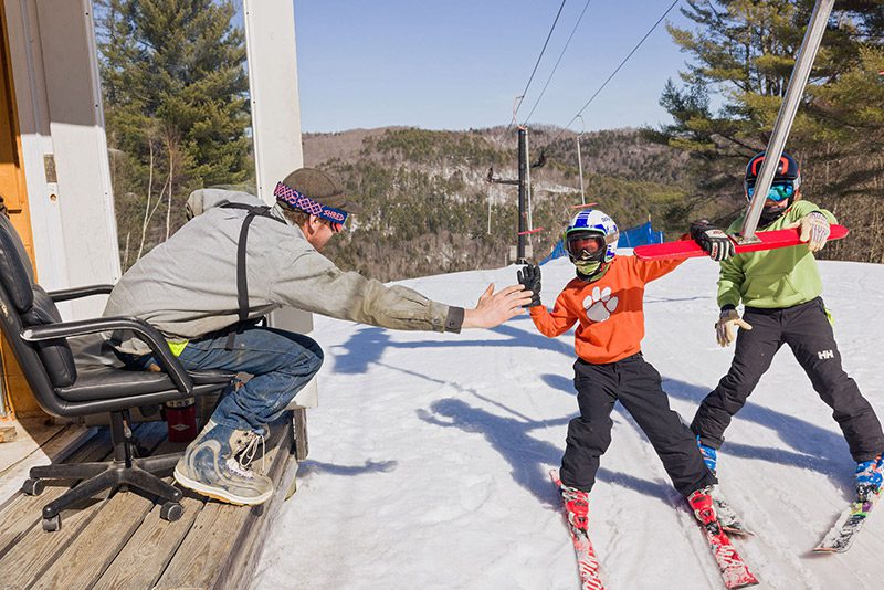 Two people ride a tow rope up a ski slope and are giving a high-five to the lift attendant at the top.