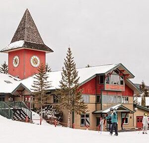 A red colored base lodge of a ski resort is behind skiers carrying skis.