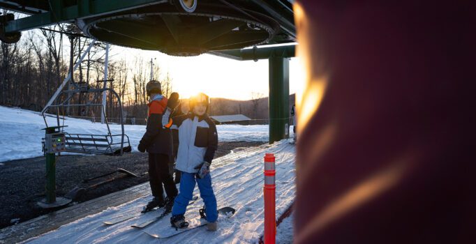 Two people stand on their snowboards waiting to take a chairlift up a ski slope.