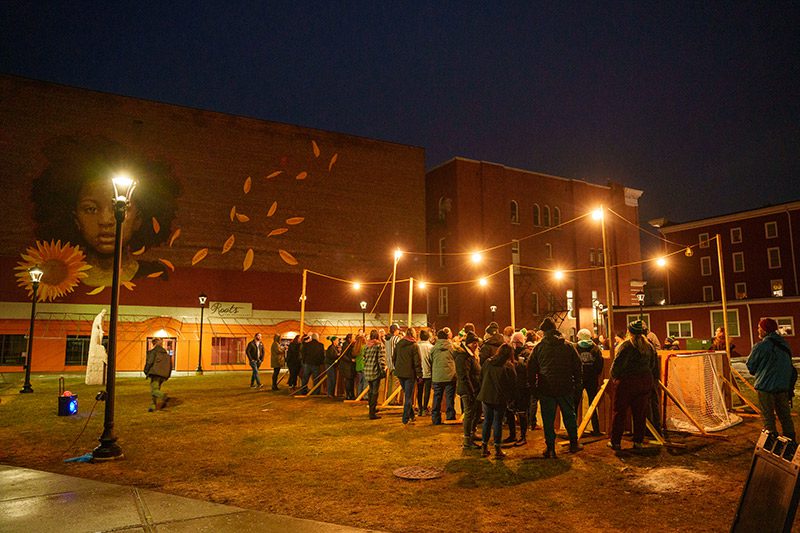 A group of people stand around a small outdoor court with wooden sides and nets at the end for a human foosball game.