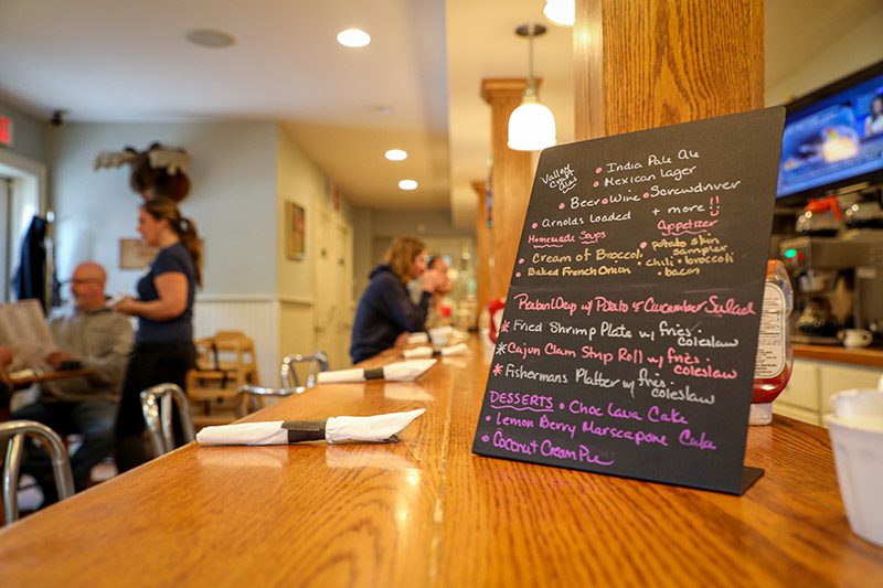 A restaurant menu sits on the bar in a small diner with patrons in the background.