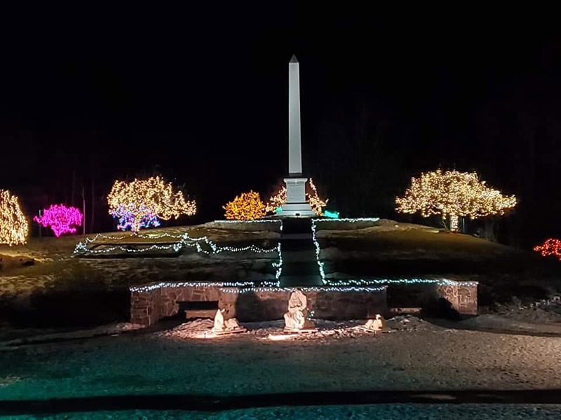 A stone obelisk is lit up at night with trees adjacent decorated with Christmas lights.