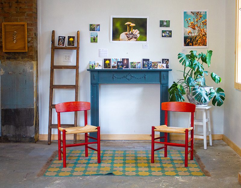 Two red chairs sit in front of an art exhibit encompassing a number of pictures on the wall and a blue fire hearth.