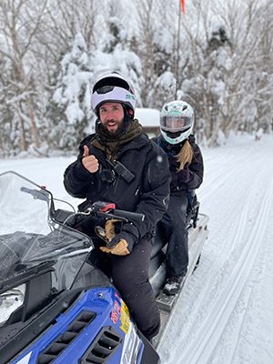 Two people sit on a snowmobile with the person in front giving a thumbs-up.