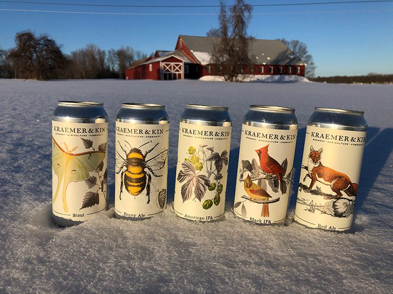 Five beer cans sit in the snow with a red barn in the background.