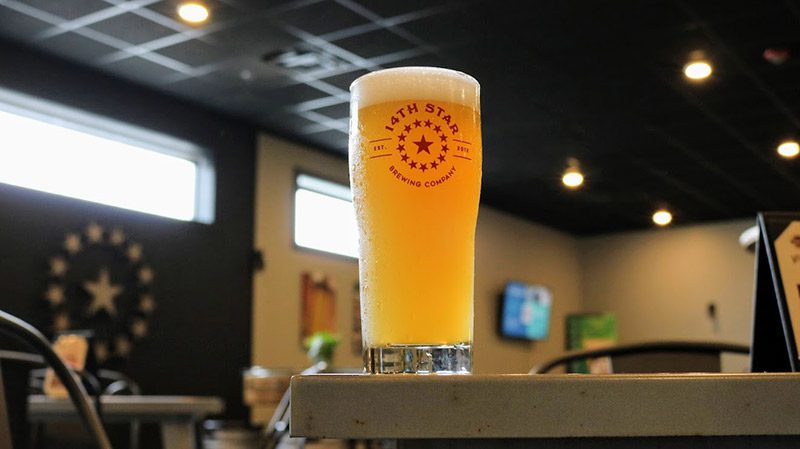 A beer glass with 14th Star Brewing Company on the side, sits on the bar of a brewery.