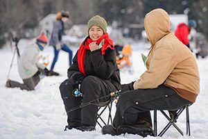 Two people hold ice fishing rods while sitting in chairs over holes in the ice.