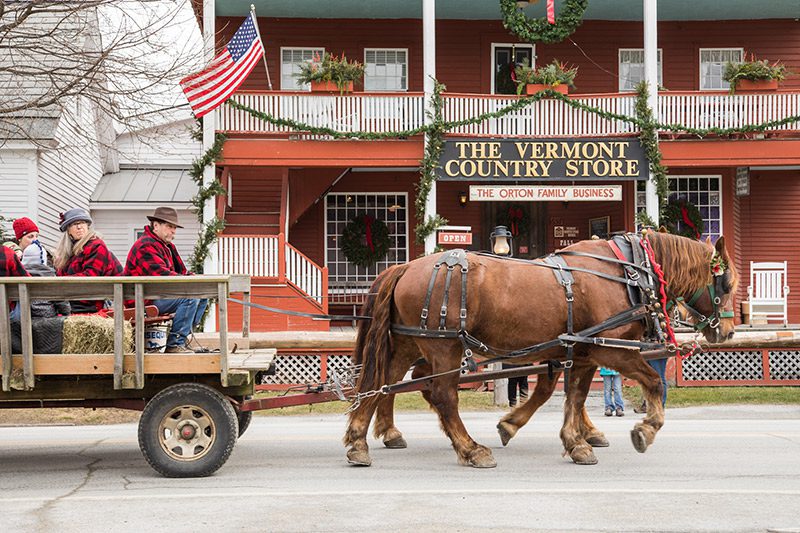 A horse pulls a trailer full of people in front of a red store named The Vermont Country Store.