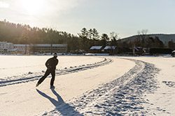 A person ice skates along a plowed path on a frozen lake at sunset.