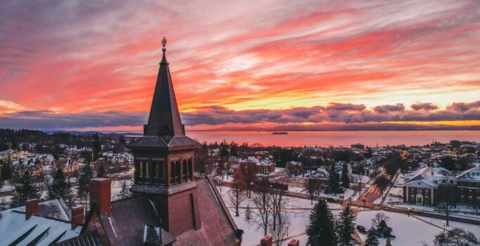 Seen from above, the sun sets in the distance across a large body of water and the buildings of a rural town in the winter.