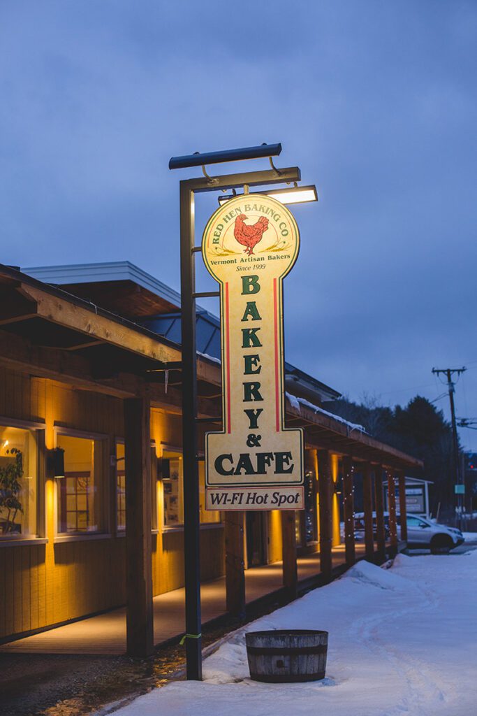 A wooden sign handing outside of a building at night in the winter that reads Red Hen Baking Co.