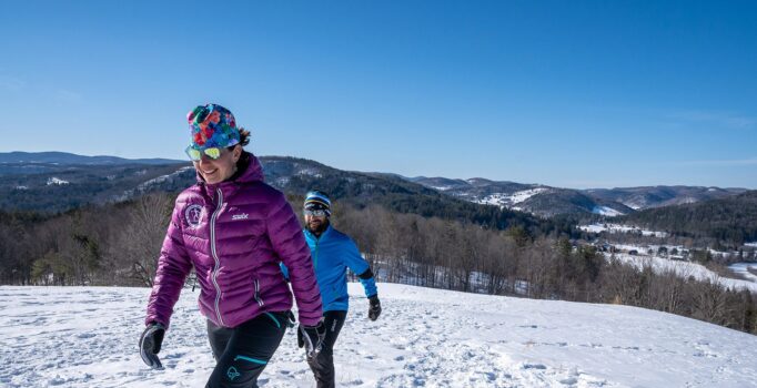 Two people snowshoe up a snowy hill with mountains in the background.