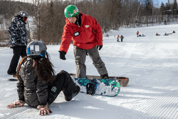 A snowboard instructor, wearing a red jacket, offers a hand to a student snowboarder who is sitting on the snow.