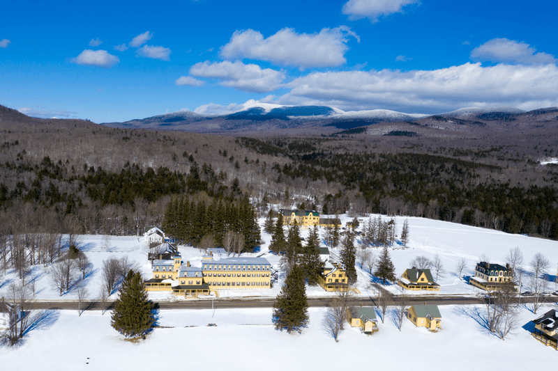 Seen from above, a wintery mountain valley and a series of yellow buildings along a country road.