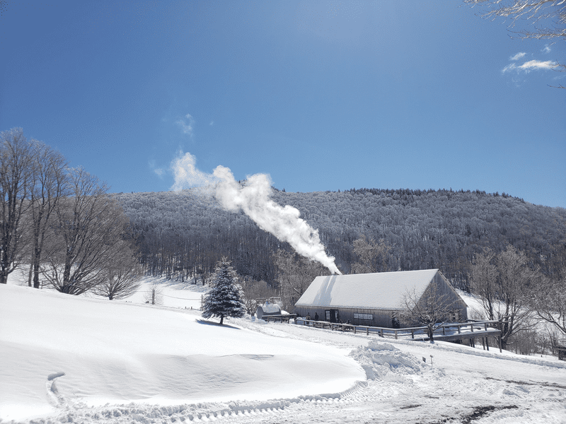 Seen from afar, a building is covered in snow and has smoke from a heating fire emitting from a smoke stack, in a snow-covered valley.