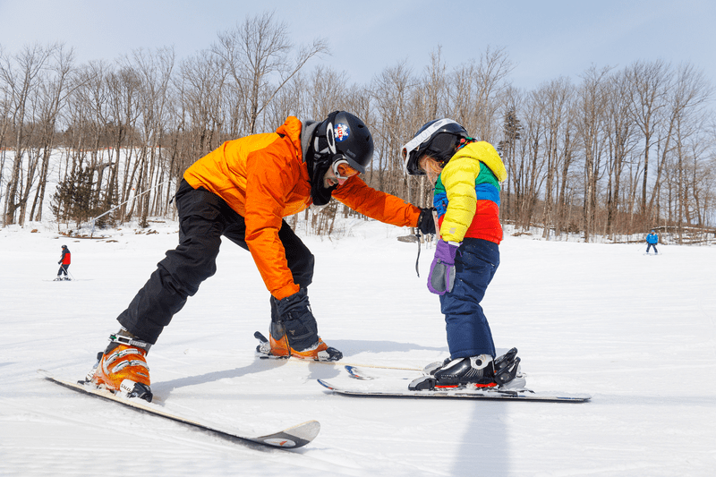 A person wearing an orange jacket teaches a child to ski at a ski resort.