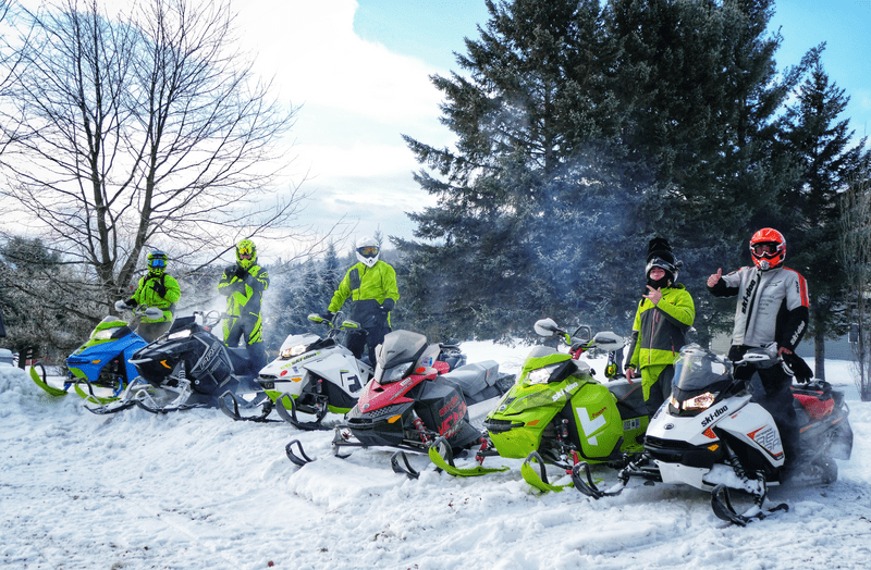 Six snowmobiles and riders stop in a clearing in the woods for a picture.