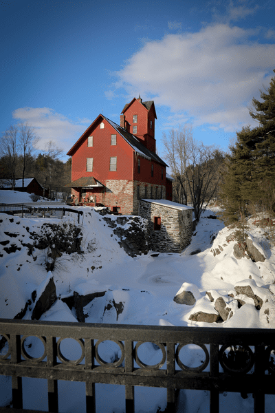 An old red mill is perched above a river and surrounded by snow.