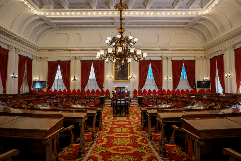 The inside of a legislative chamber, with red velvet drapes on the windows and ornate red carpets.