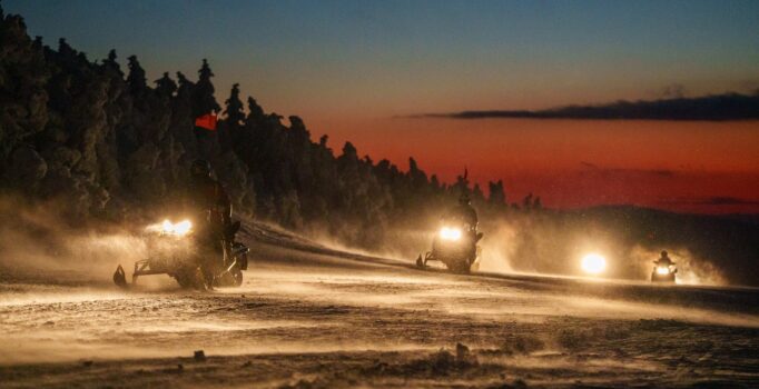 Four snowmobiles with the head lights on, ride toward the camera at sunset.