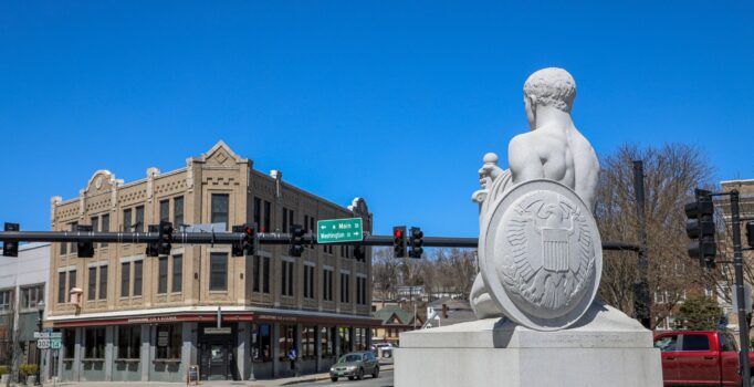 A granite sculpture of a person holding a sword and shield, faces out toward a town street and downtown buildings.