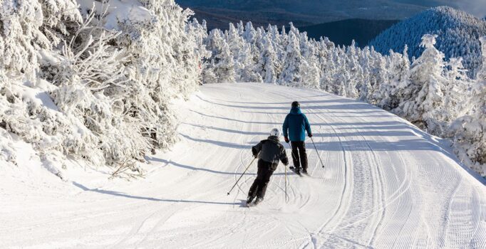 Two people ski down a snowy mountain slope on a sunny day.