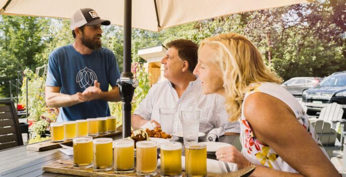A server takes a meal order from two patrons at an outdoor restaurant on a warm day.