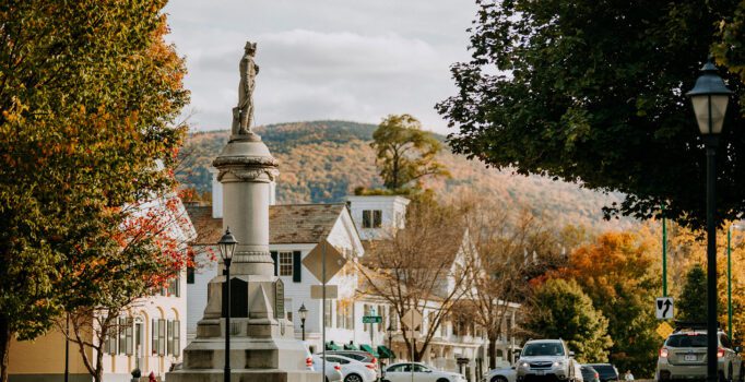 Old white buildings and a large statue are next to a rural town road in the fall.