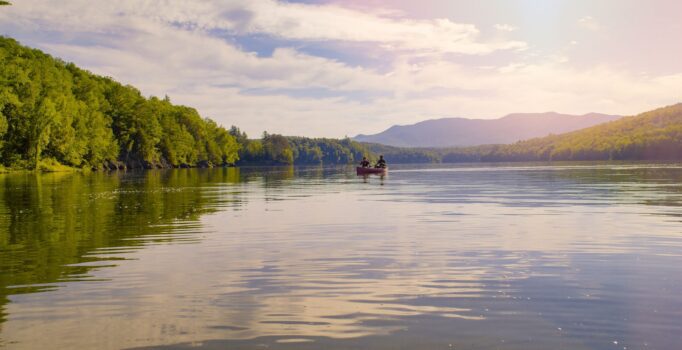 Two people canoe on a lake surrounded by forests and mountains.