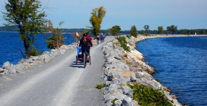 People walk or ride bikes across a gravel path on a blue lake outdoors on a sunny day.
