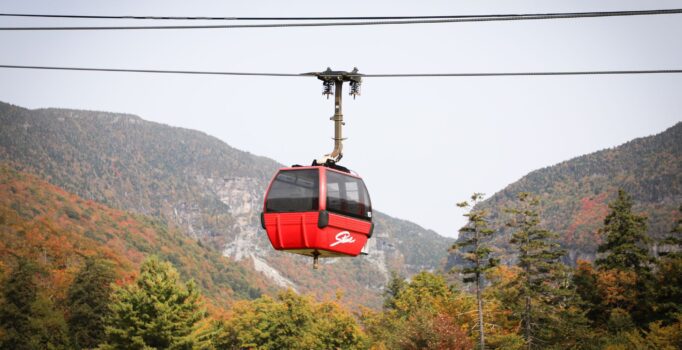A red gondola reading Stowe in stylized script moves over a mountain with fall-colored trees. 