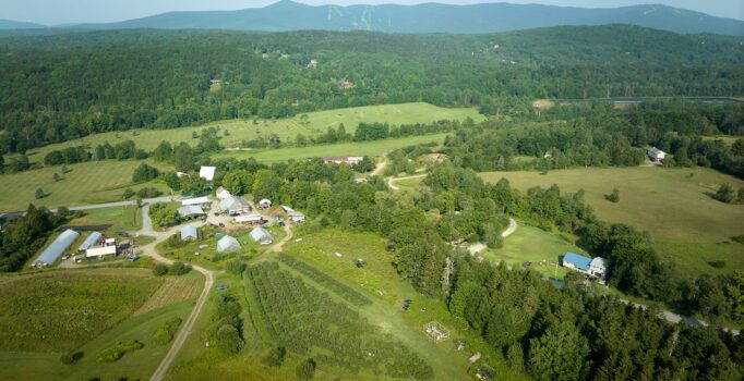 Seen from above, a small farm nestled in the trees and mountains in the background.