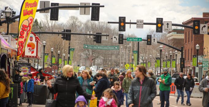 Groups of people celebrate in a street lined with festival vendors on a spring day.