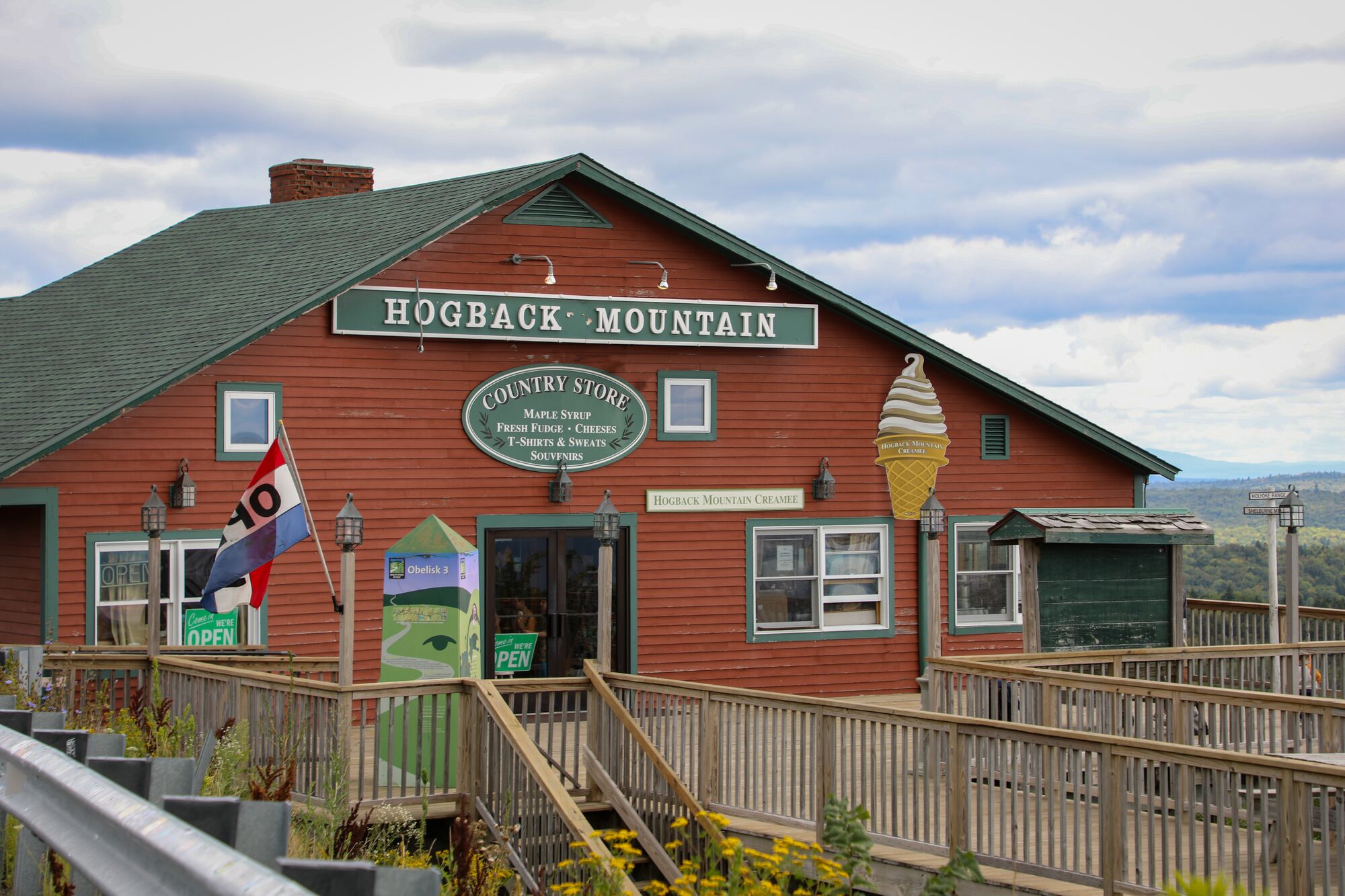  A red general store building seen from outside in the summer with grass growing and flowers blooming. Behind the building, mountains are visible.