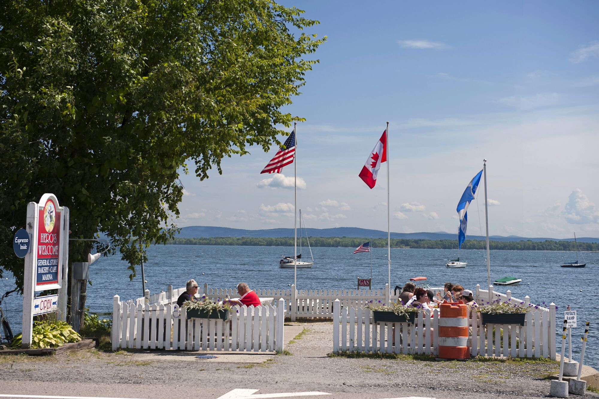 Various flags fly overhead on a summer day as people dine by a lake. 