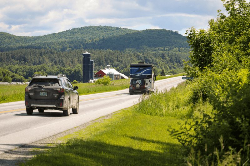 Cars on a road in the summer. A barn is visible behind them on the left.