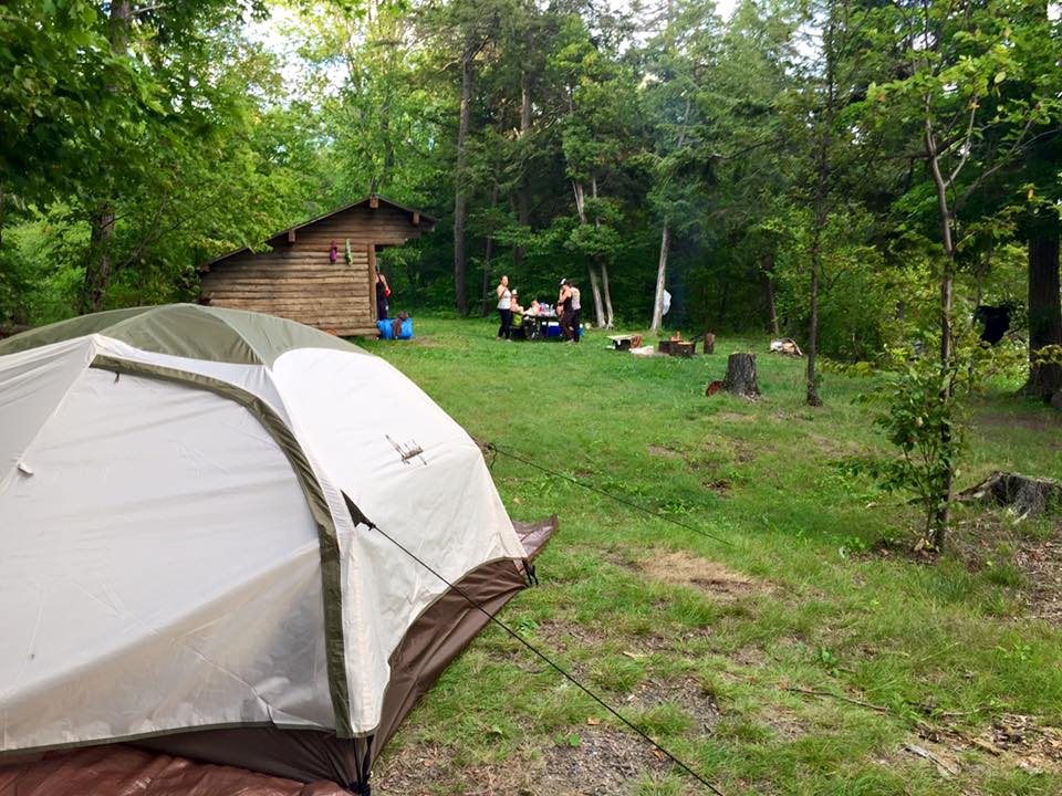 A tent is pitched in a grassy campsite surrounded by dense trees. Nearby, people gather around a picnic table. A rustic wooden cabin is in the background.