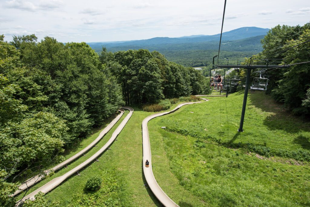 A scenic view of a summer mountain slide winding through greenery, with a chairlift carrying riders above.
