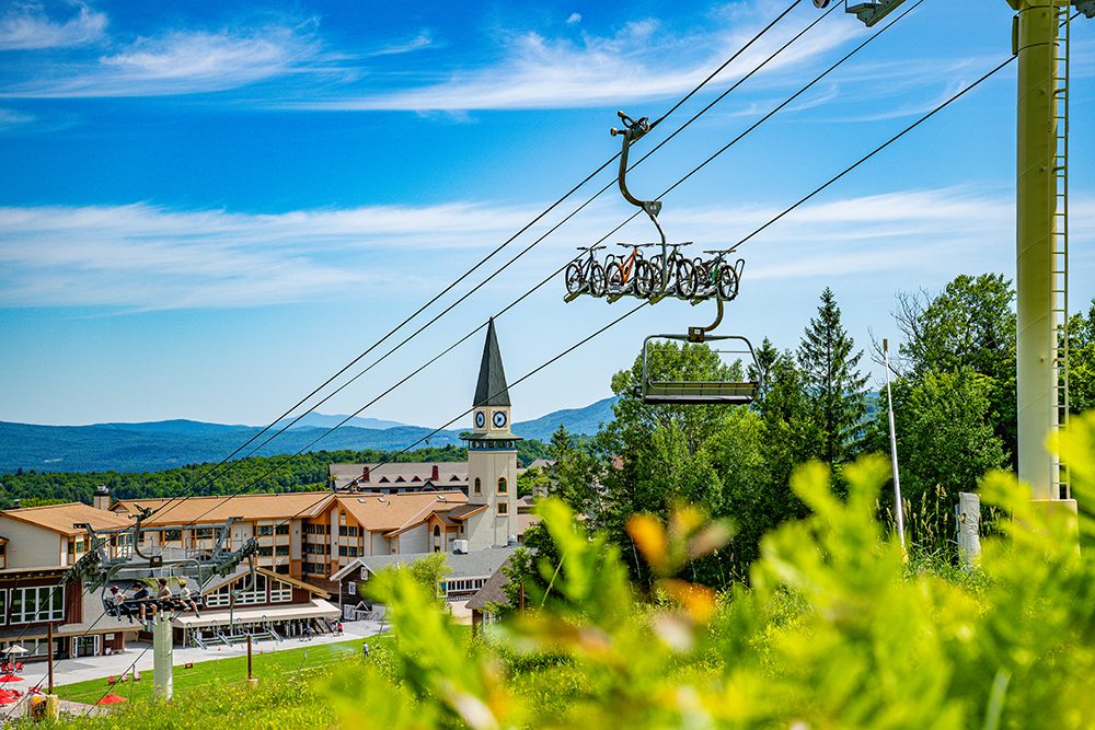 A ski resort with a chairlift carrying mountain bikes, set against a backdrop of a clock tower, buildings, green trees, and distant mountains under a blue sky.