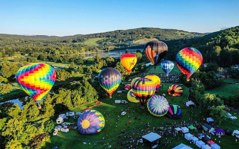 Aerial view of a vibrant hot air balloon festival with colorful balloons floating above a lush green landscape and excited crowds below.