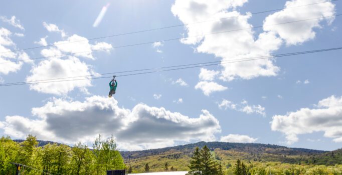 Person ziplining through sunny sky with fluffy clouds, over a lush green landscape with hills and trees.