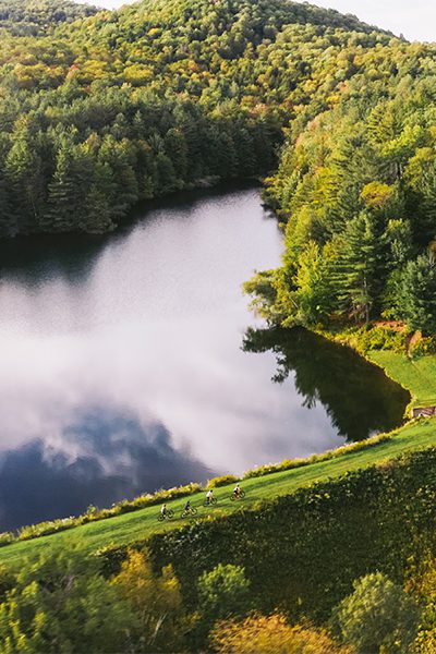 A serene landscape featuring a reflective lake surrounded by vibrant trees, with a grassy hillside and cyclists in the distance.