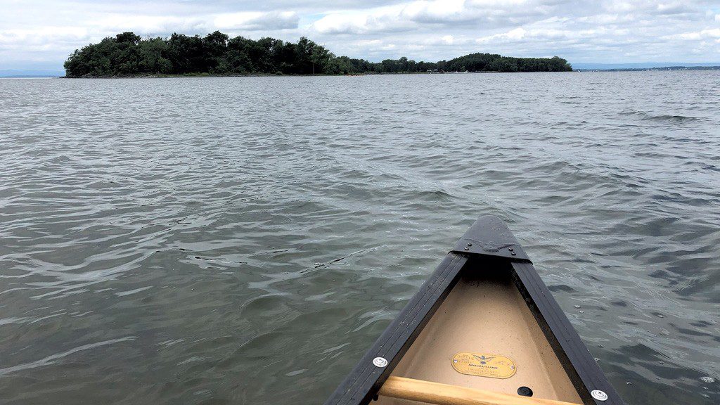 A canoe glides through calm gray waters toward a tree-covered island under a cloudy sky, evoking a serene and adventurous atmosphere.