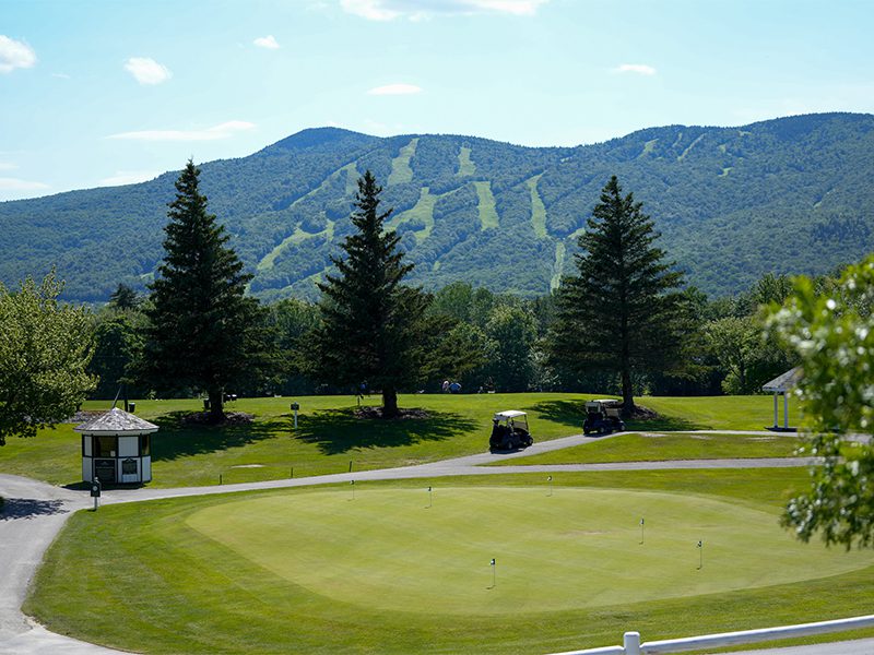 A golf course with a putting green, golf carts, and a small building in the foreground; tree-covered mountains with ski trails in the background under a clear sky.