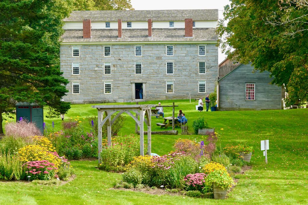 A large stone building with white trim stands behind a garden filled with colorful flowers, with several people walking and sitting on the lawn.