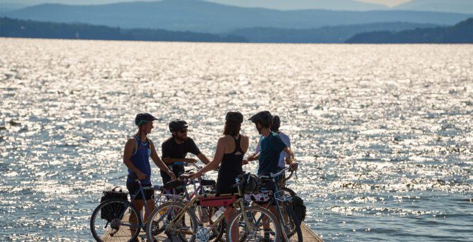 Five people with bicycles stand and talk on a dock overlooking a sunlit body of water with mountains in the background.